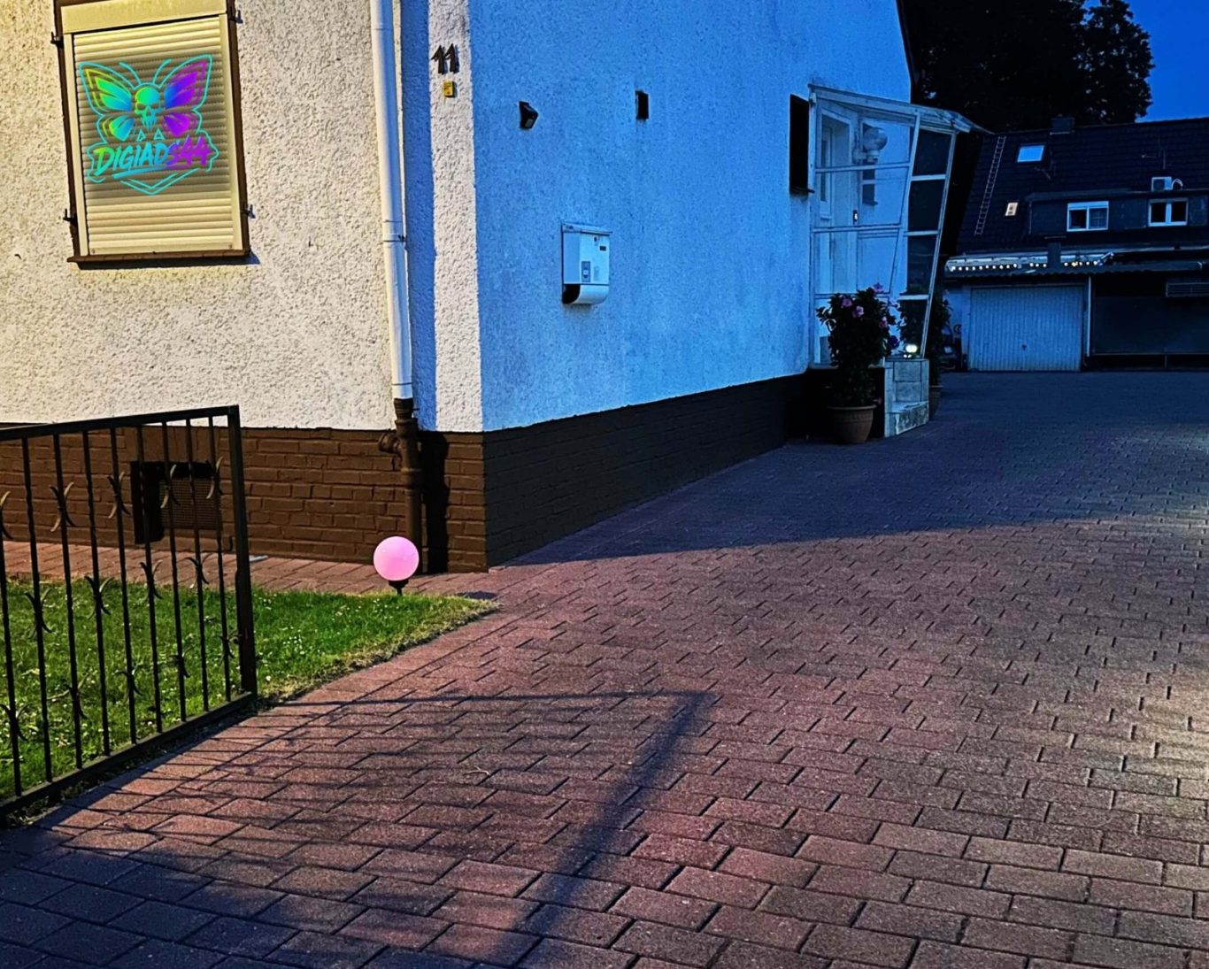 DigiAds44: Digitale Präsenz auch bei Nacht Abendansicht des Hauses DigiAds44 mit beleuchtetem Fensterlogo und gepflasterter Einfahrt, rosa Leuchte im Vorgarten